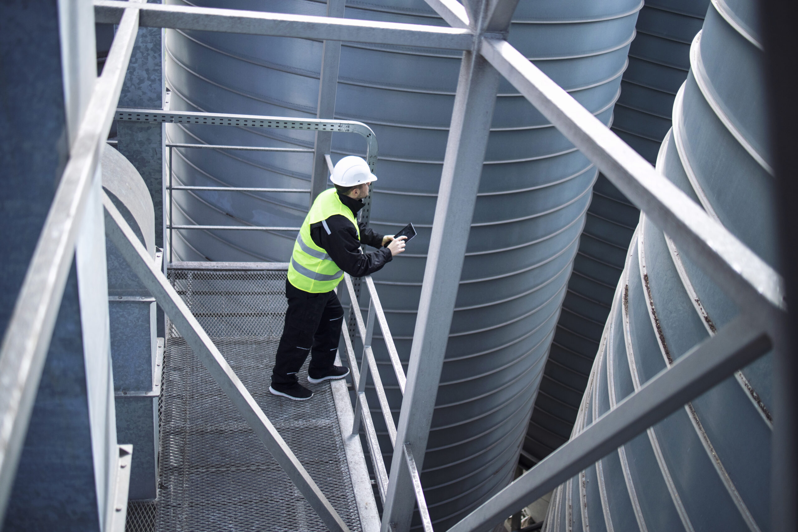 Factory silos worker standing on metal platform between industrial storage tanks and looking at tablet about food production.